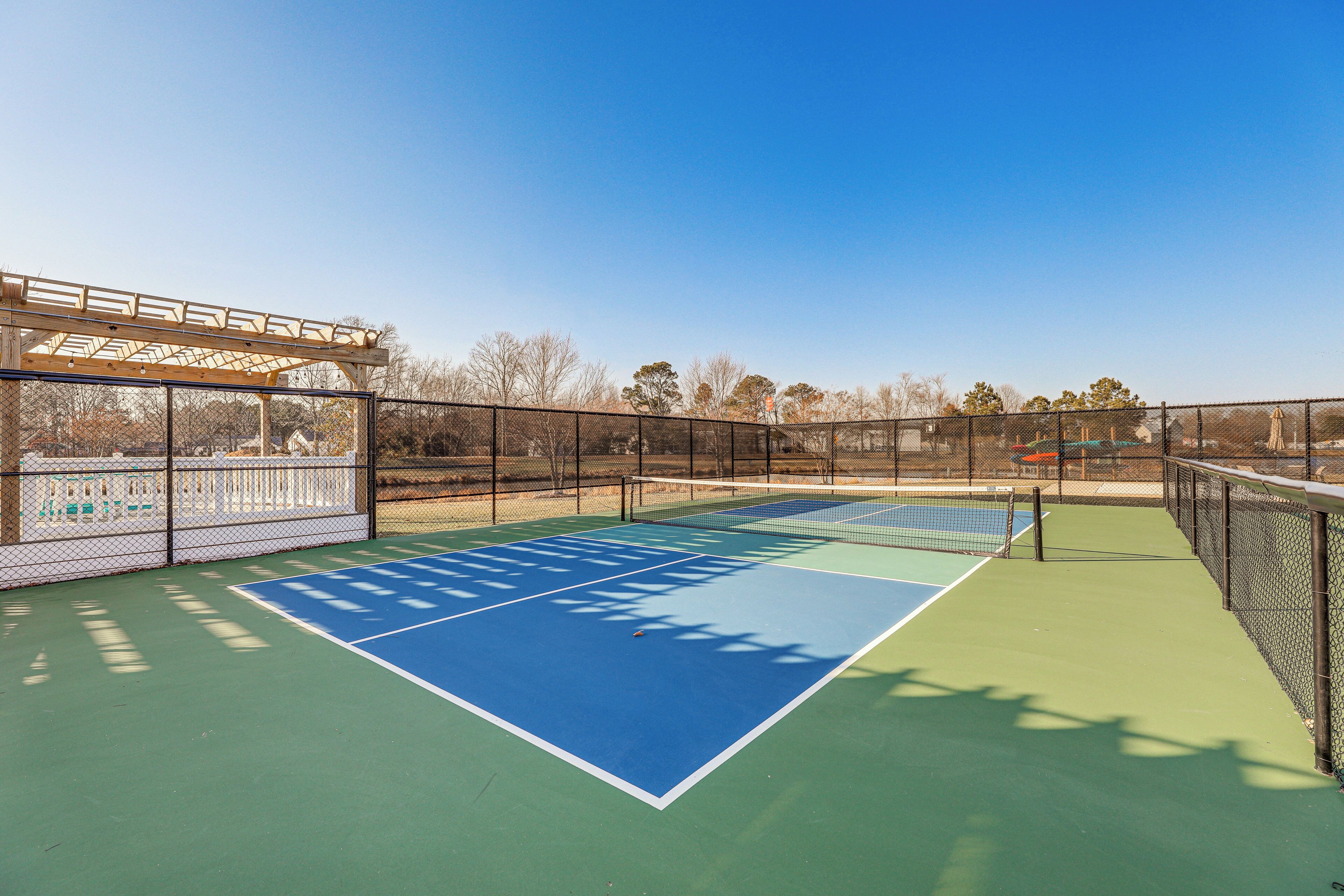 A tennis court with a net, a wooden pergola, and a fence.