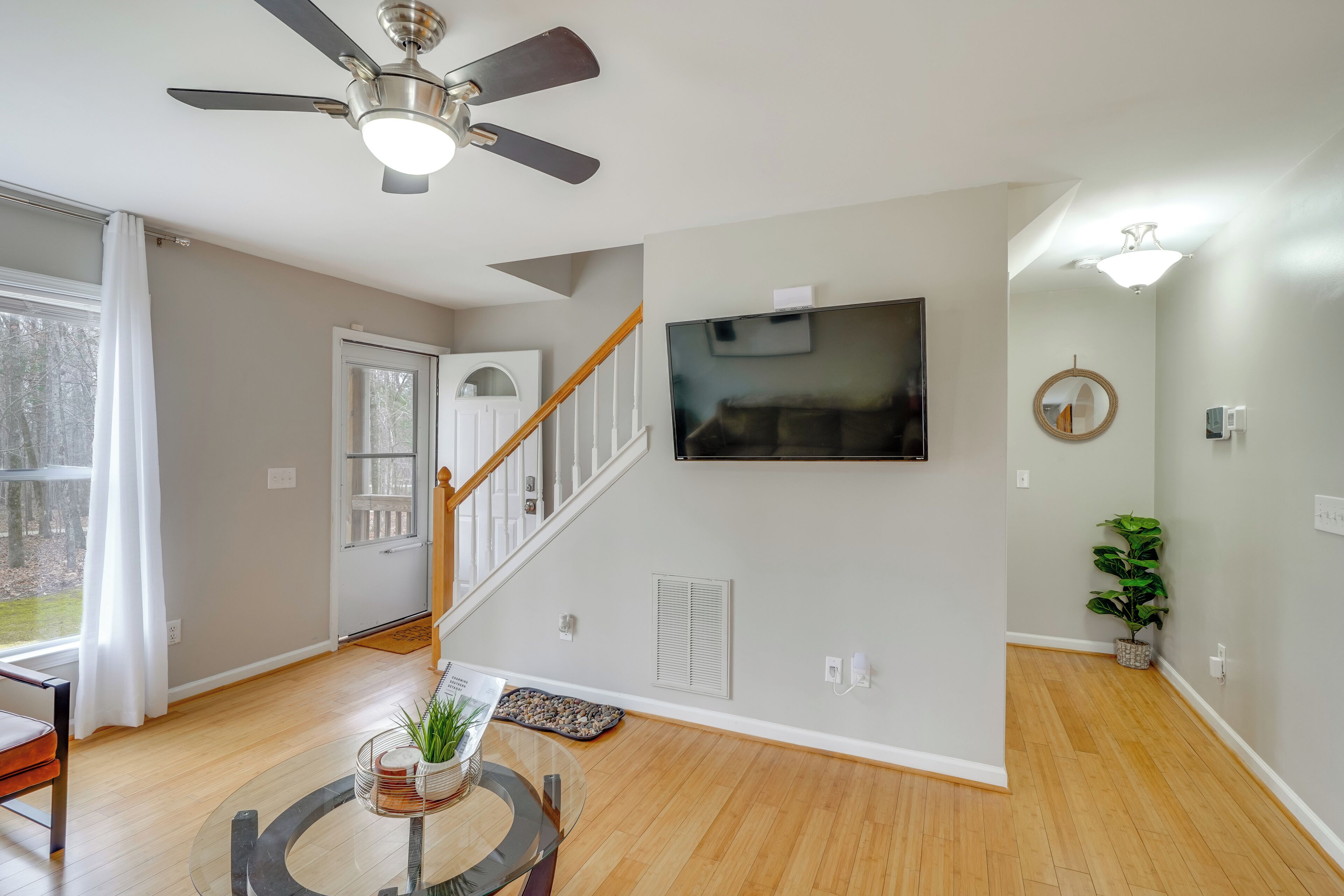 A modern living room with a wooden floor, a ceiling fan, a staircase, a flat-screen TV, and a potted plant.