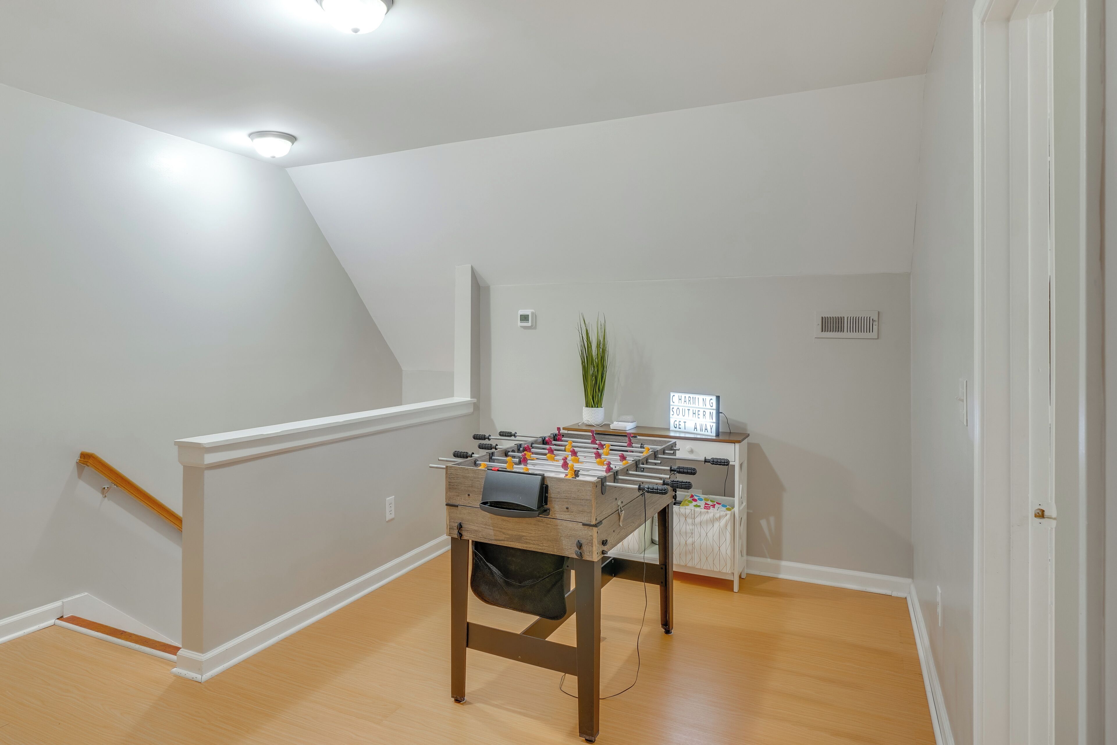 A room with a foosball table, a desk with a computer, a plant, and a sign that says 'CHARMING SOUTHERN N GET AWAY'.