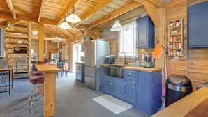 A rustic cabin interior with a wooden ceiling, a kitchen area with blue cabinets, a bar stool, and a dining table. - Private Hot Tub! Country Retreat in West Virginia (Friendly)