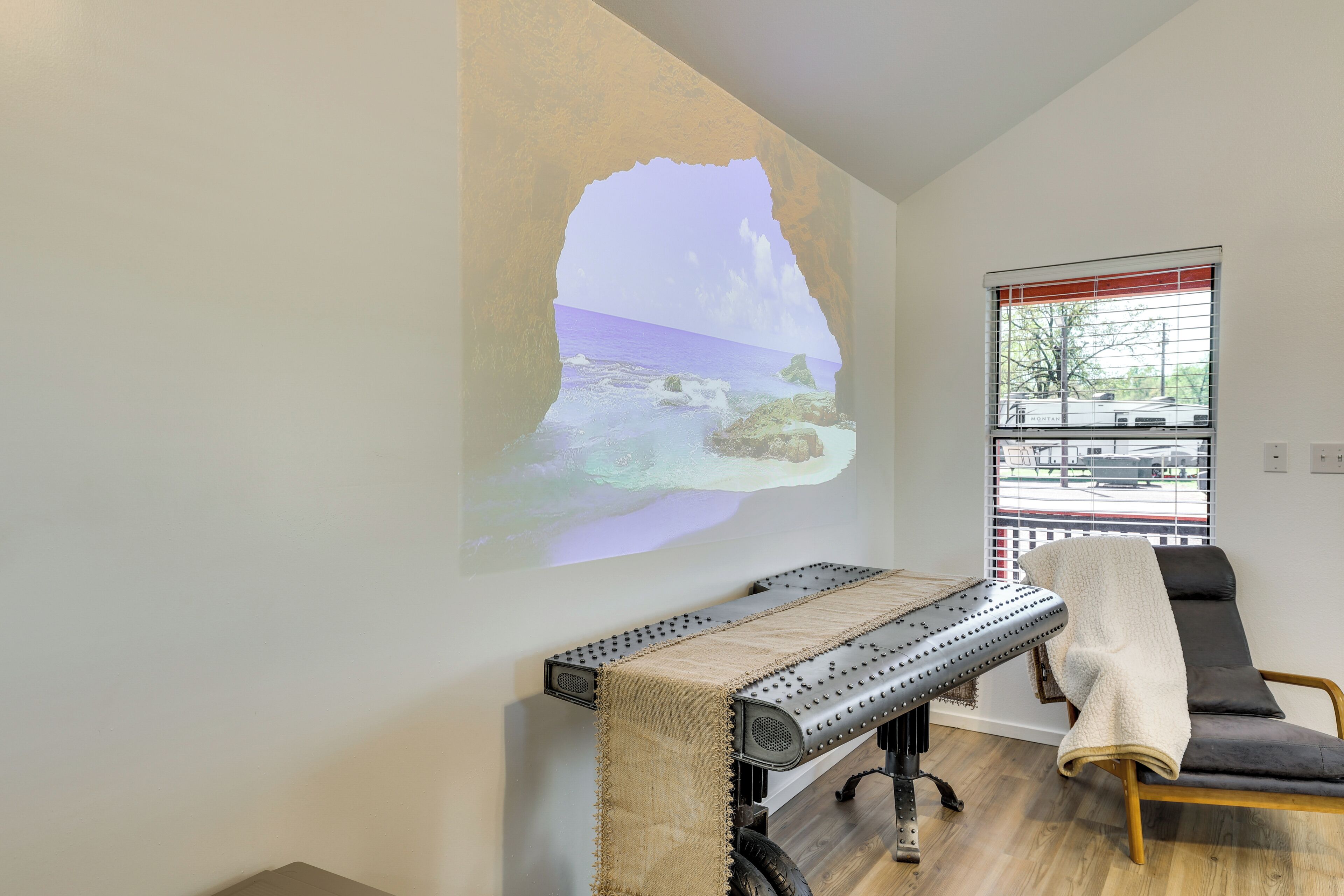 A room with a projection screen displaying a beach scene, a massage table, a window with blinds, and a chair with a blanket.