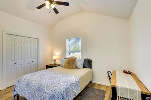Une chambre à coucher comprenant un lit, une table de chevet, une lampe, un ventilateur de plafond, une fenêtre avec des stores, une porte et un banc recouvert d’une nappe.