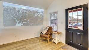 A room with a projection screen displaying a mountainous landscape, a wooden chair, a small table with a bowl, and a dark door with a window. - Piney Woods Tiny Cabin: 3 Mi to Sabine River! (Joaquin)