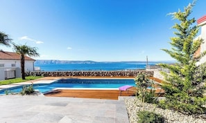 A swimming pool with a wooden deck, a stone wall, and a view of the sea. - Modern apartment with sea view & pool in Klenovica (Klenovica)