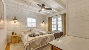 Ein Schlafzimmer mit Bett, Nachttischlampen, Deckenventilator, einem Fenster mit Vorhängen und einem Holztisch.