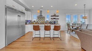 Eine moderne Küche mit Edelstahlkühlschrank, Holzbarhocker, Pendelleuchten und einem Essbereich mit Sofa und Tisch.