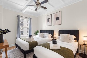 Une chambre d’hôtel avec deux lits, un ventilateur de plafond, une fenêtre avec des rideaux et des œuvres d’art encadrées accrochées au mur.