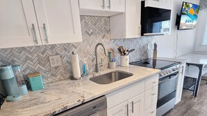 Una cocina con gabinetes blancos, salpicadero con motivo de espiga, un lavamanos de acero inoxidable, una cafetera y una estufa.