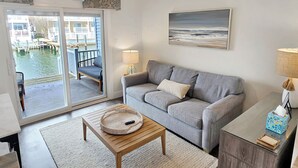Una sala de estar con un sofá gris, una mesa de centro, una lámpara, un cuadro y vista a un cuerpo de agua a través de una puerta corredera.