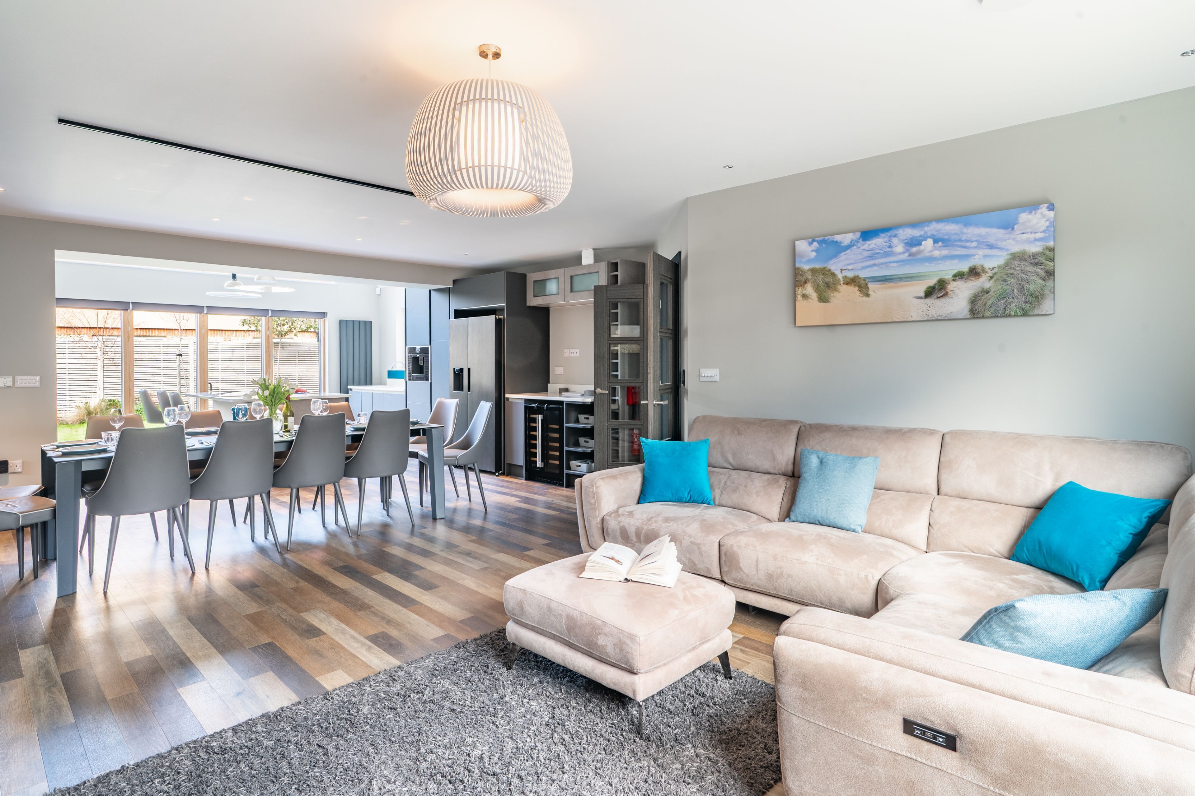 Ein modernes Wohnzimmer mit einem beigefarbenen Sofa, einem Essbereich mit Stühlen, einer Küche im Hintergrund und einem großen Fenster.