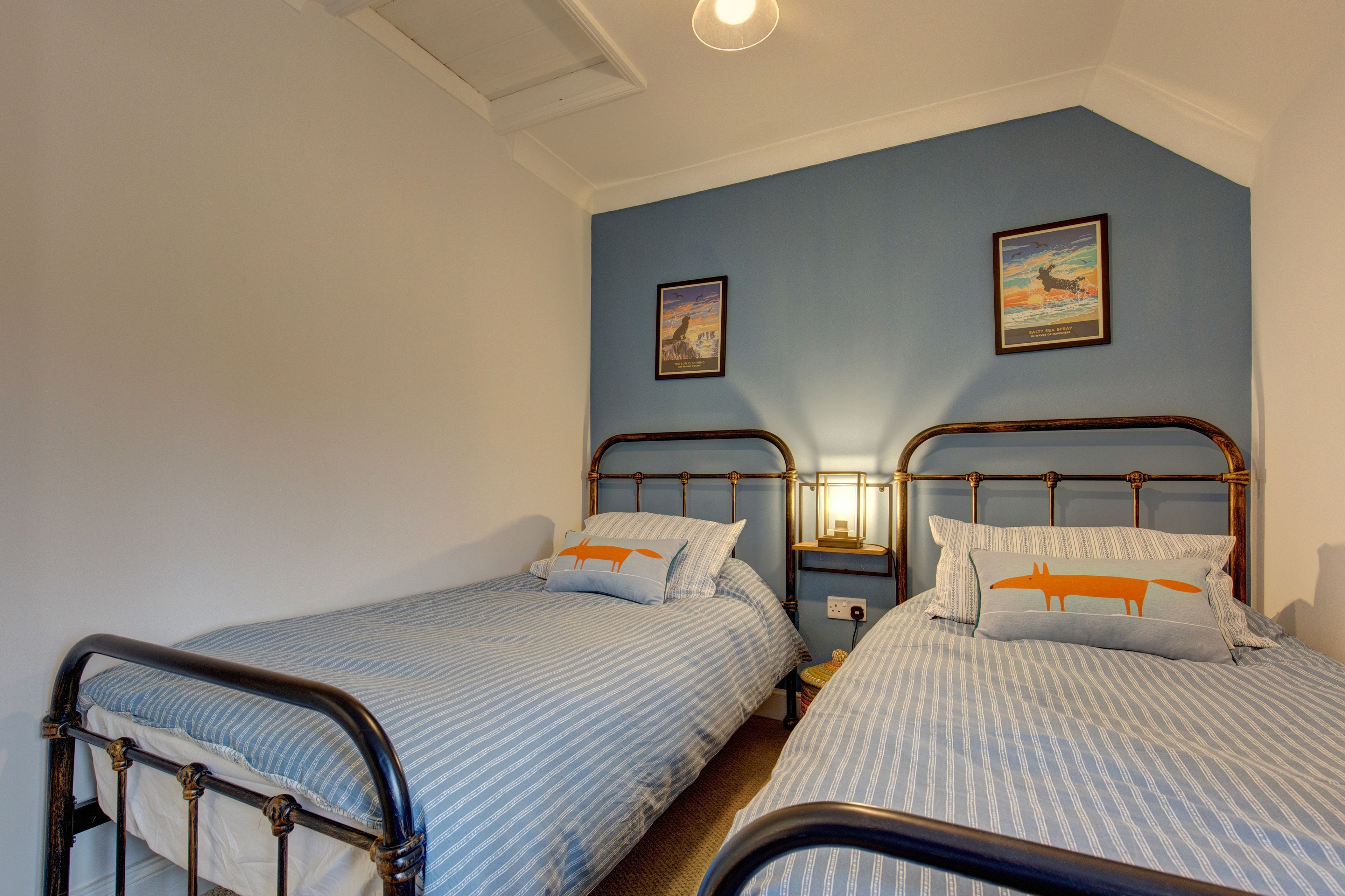 A bedroom with two single beds, a nightstand with a lamp, and two framed pictures on the wall.