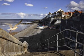 A coastal scene with a staircase leading down to the beach, a seagull flying, and boats on the shore. - 1 The Cottages, Aylmerton, Norfolk (Aylmerton)