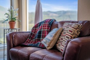A brown leather sofa with plaid and patterned cushions, a potted plant, and a view of mountains through a window.