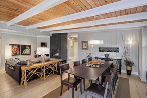 Ein modernes Wohnzimmer mit Holzbalkendecke, Kamin, Essbereich mit großem Tisch und Stühlen sowie einer Couch mit dekorativem Konsolentisch.
