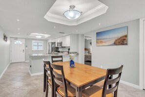 A dining area with a wooden table and chairs, a kitchen in the background, and a beach-themed painting on the wall. - 🏖️Across the Street from Beach - Quick Walk!🐬Heated Pool🌴Madeira Place 201 (Madeira Beach)