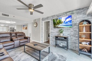 Ein Wohnzimmer mit einer Ledersofa, einem Holztisch, einem an einer Steinwand montierten Fernseher, einem Deckenventilator und einem dekorativen Regal mit einem Seestern.