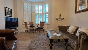 A living room with a brown leather sofa, a glass coffee table, wicker chairs, a television, and a bay window with shutters. - 2-bed Garden Flat in Fulham (London)