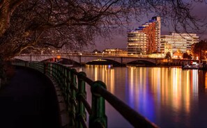 A nighttime view of a cityscape with a river, a bridge, and modern buildings with illuminated windows. - 2-bed Garden Flat in Fulham (London)