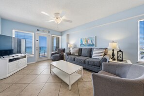 A living room with a television, a ceiling fan, a sofa, a coffee table, and a painting on the wall. - Beachfront Corner Condo | Gulf Shores | Sleeps 6 | Pool & On-Site Dining (Gulf Shores)