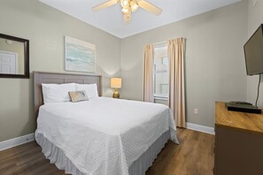 Une chambre à coucher bien rangée, avec un lit, une lampe de chevet, une fenêtre avec des rideaux, un ventilateur de plafond et un téléviseur posé sur un meuble en bois.