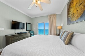 A hotel room with a large bed, a ceiling fan, a TV, a dresser, a balcony with curtains, and a painting. - Gulf Shores Condo | Direct Beach Access | Sleeps 6 (Gulf Shores)