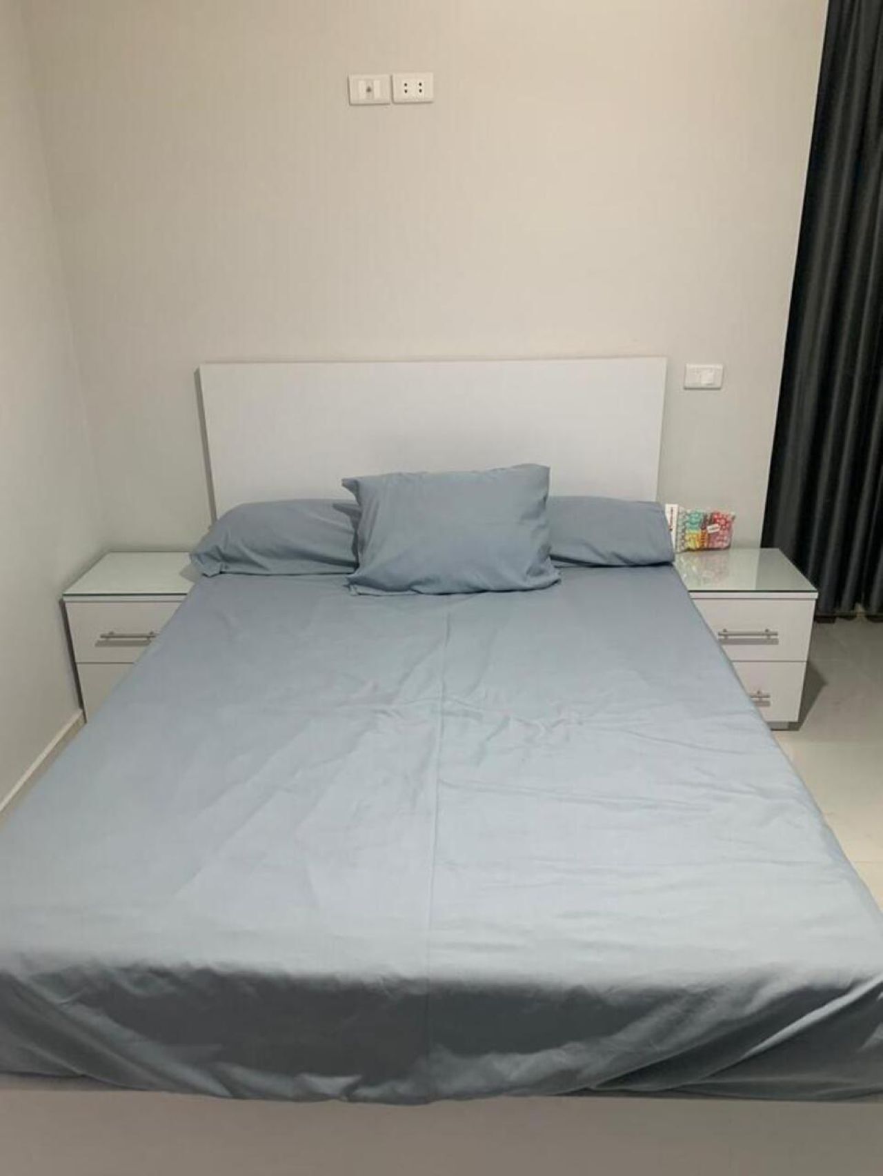 Una cama con colchón y almohadas grises, dos mesitas de noche con cajones, y un enchufe en la pared.