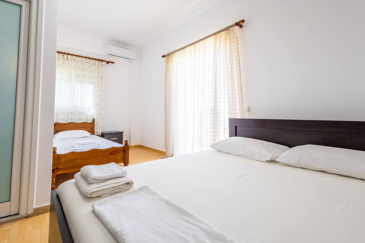Une chambre à coucher avec un lit en bois, du linge de lit blanc, une table de chevet et une fenêtre avec des rideaux.