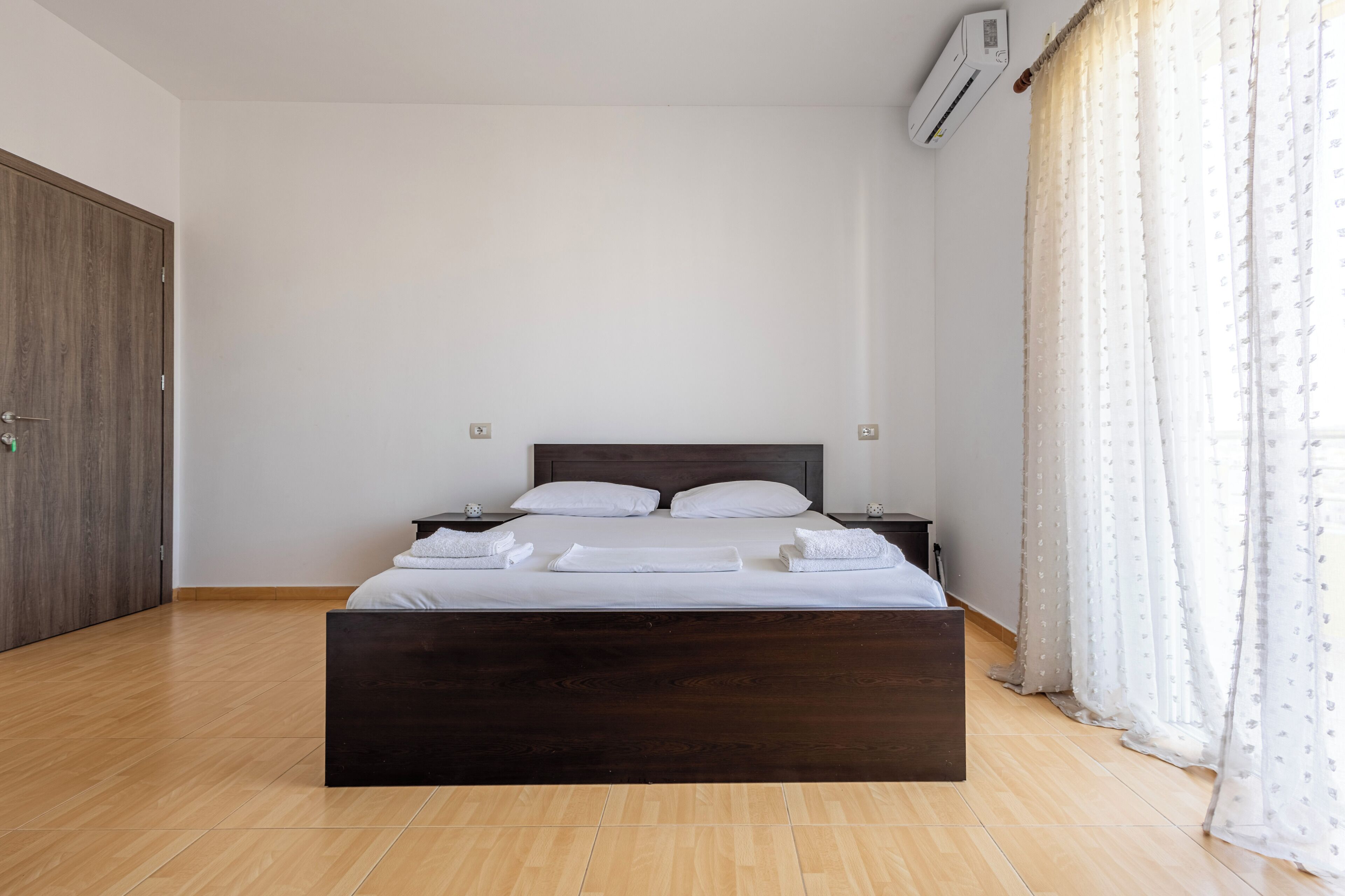 Une chambre à coucher avec un lit en bois, du linge de lit blanc, une table de chevet, une armoire et une fenêtre ornée de rideaux légers.