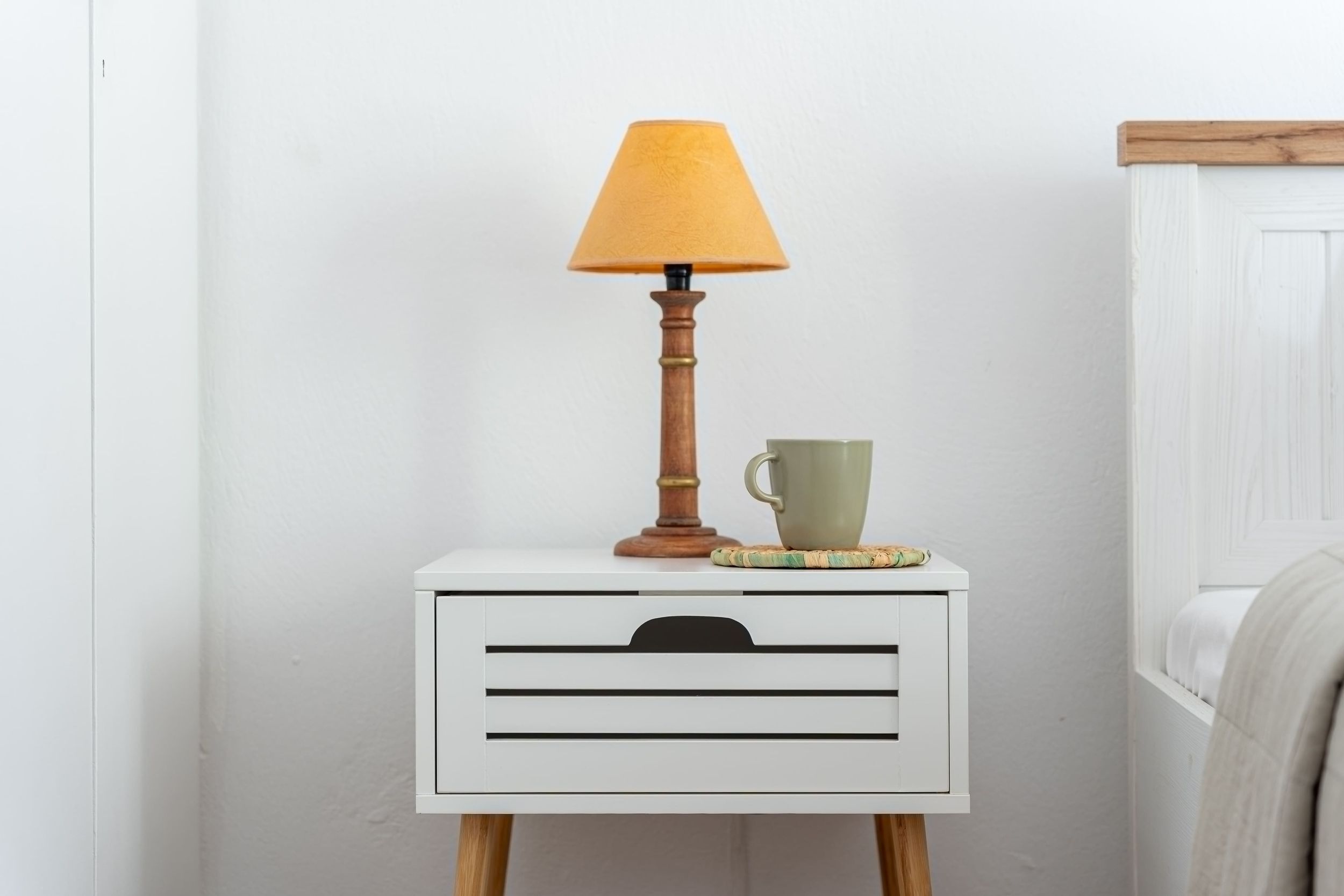 Een witte nachtkastje met een houten lamp, een groene mok en een onderzetter.