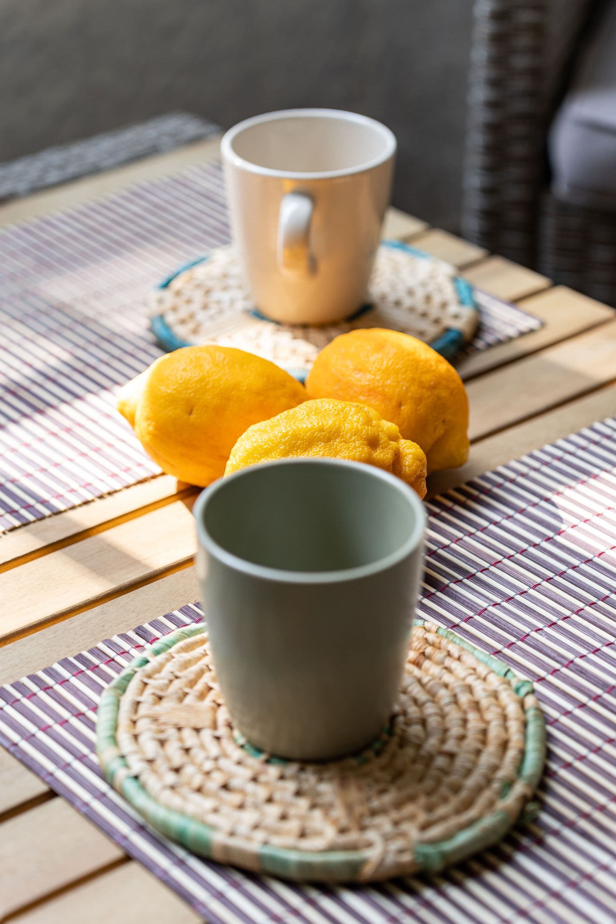 A table with a woven placemat, a grey mug, and two lemons.