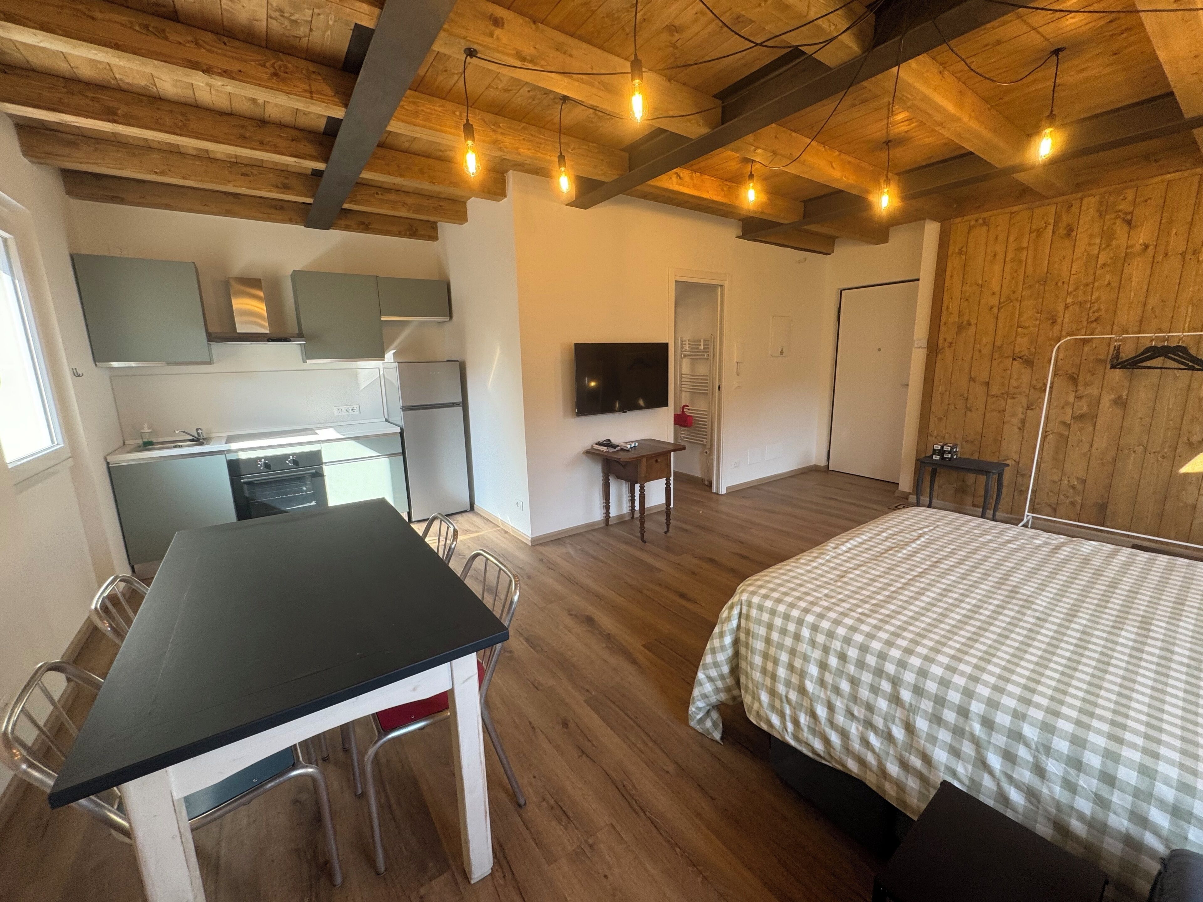 A compact room with a kitchenette, a dining area, a bed, and a TV mounted on the wall.