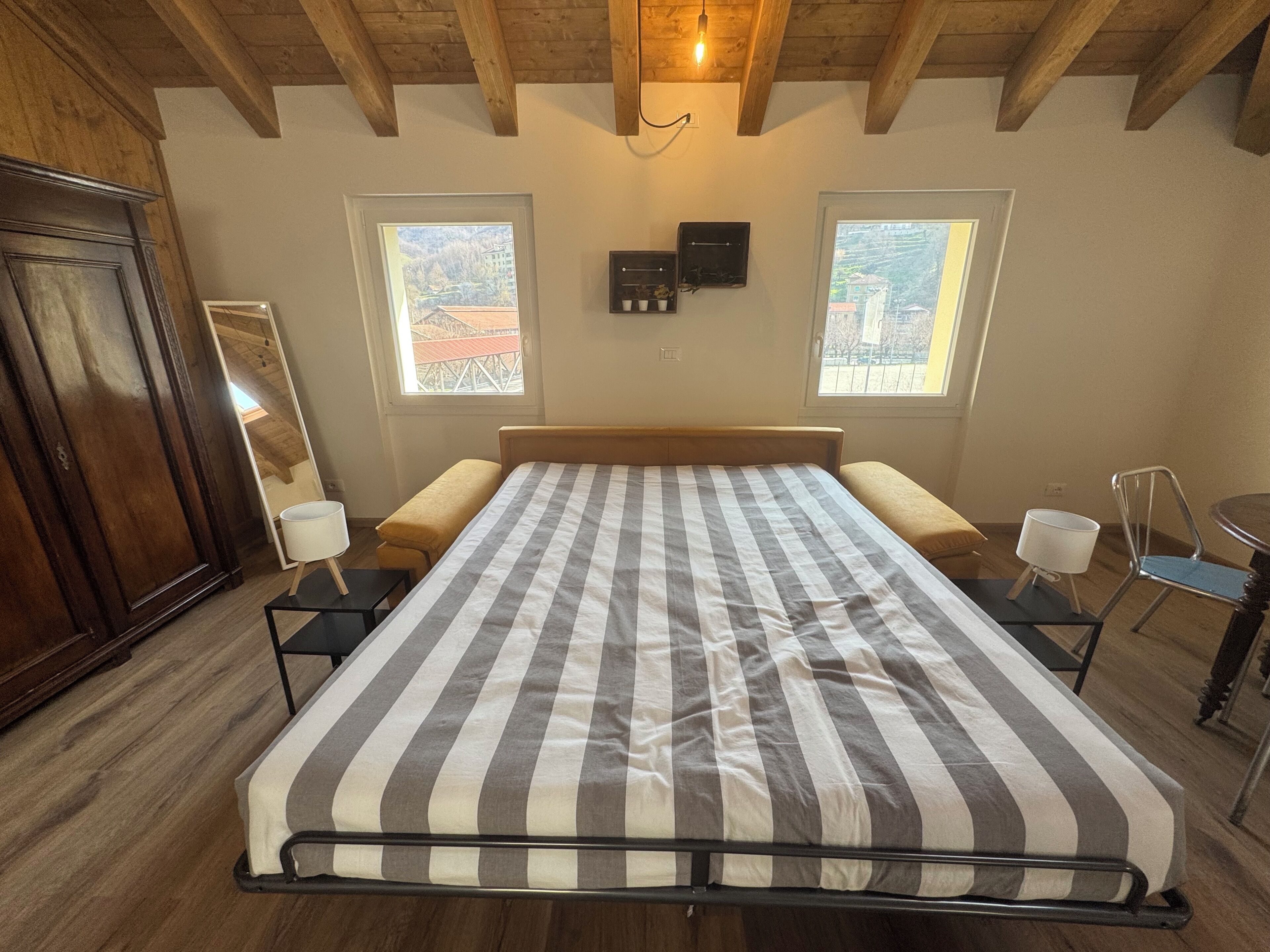 A bedroom with a bed, two bedside tables, a wooden wardrobe, and two windows with a view.