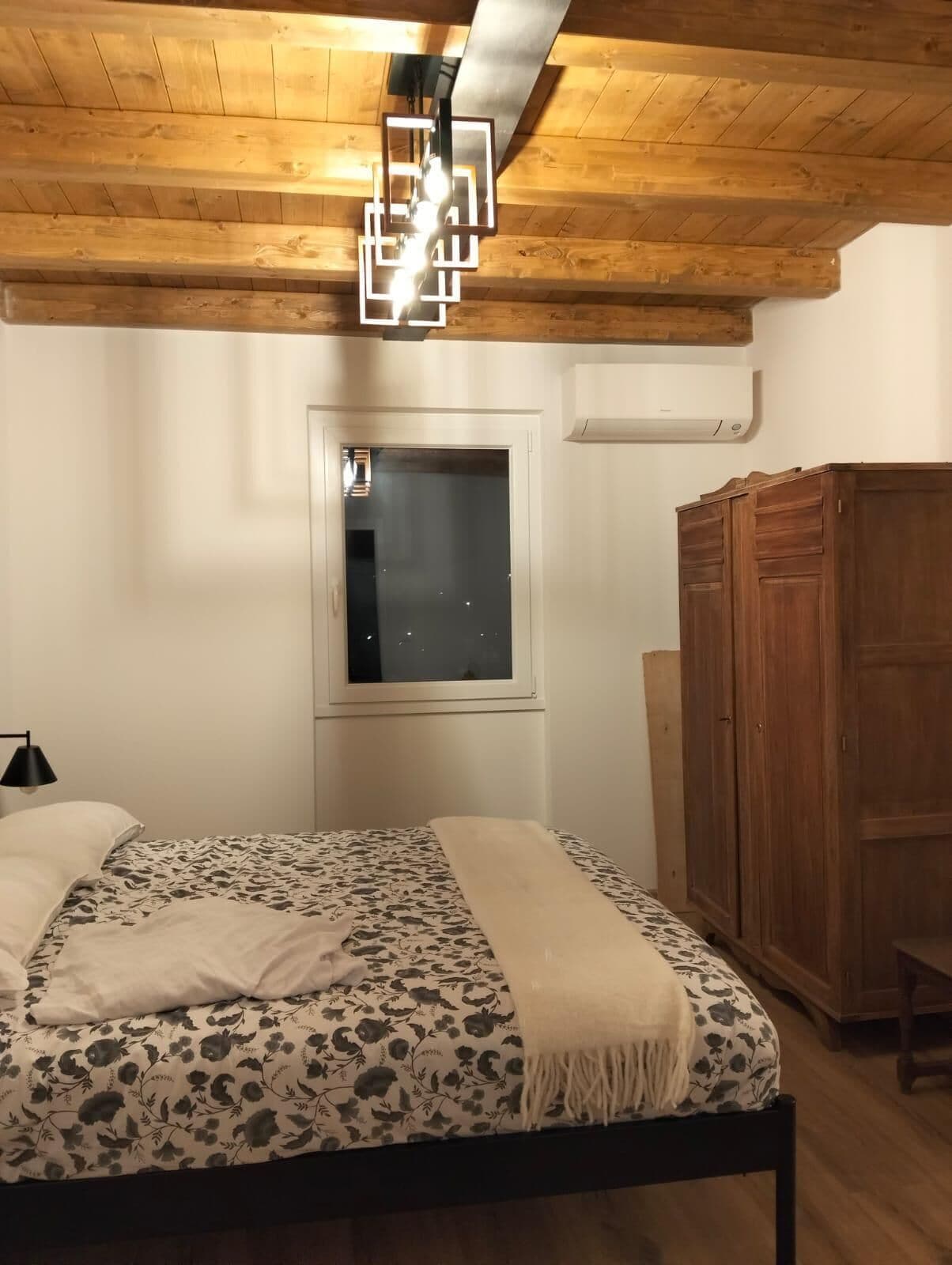 A bedroom with a wooden ceiling, a bed with floral bedding, a window, and a wardrobe.