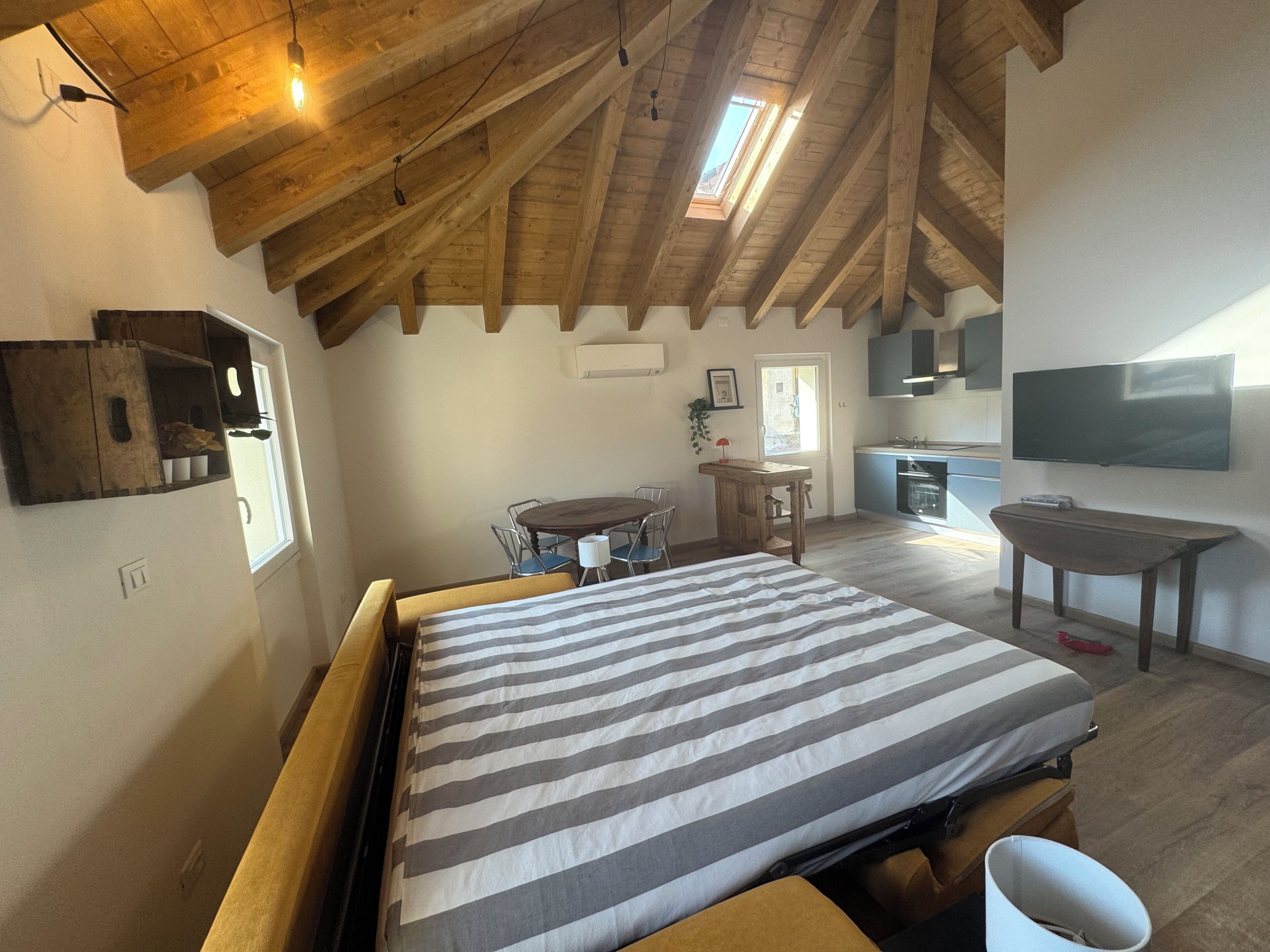 A bedroom with a bed, a wooden ceiling, a window, a dining table, chairs, a small kitchen area, and a TV mounted on the wall.