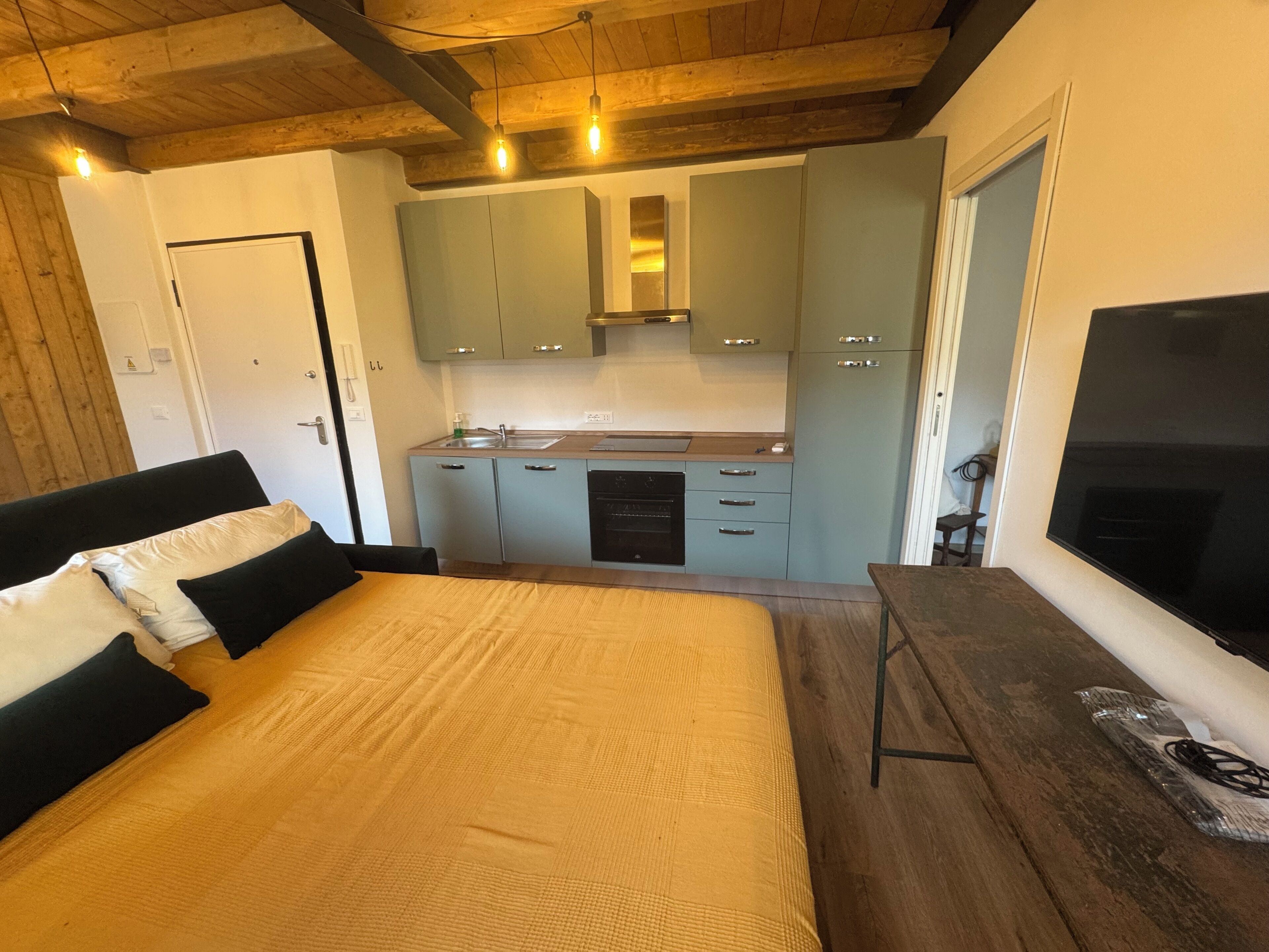 Un dormitorio con cama, techo de madera, cocina con armarios azul claro y televisor.