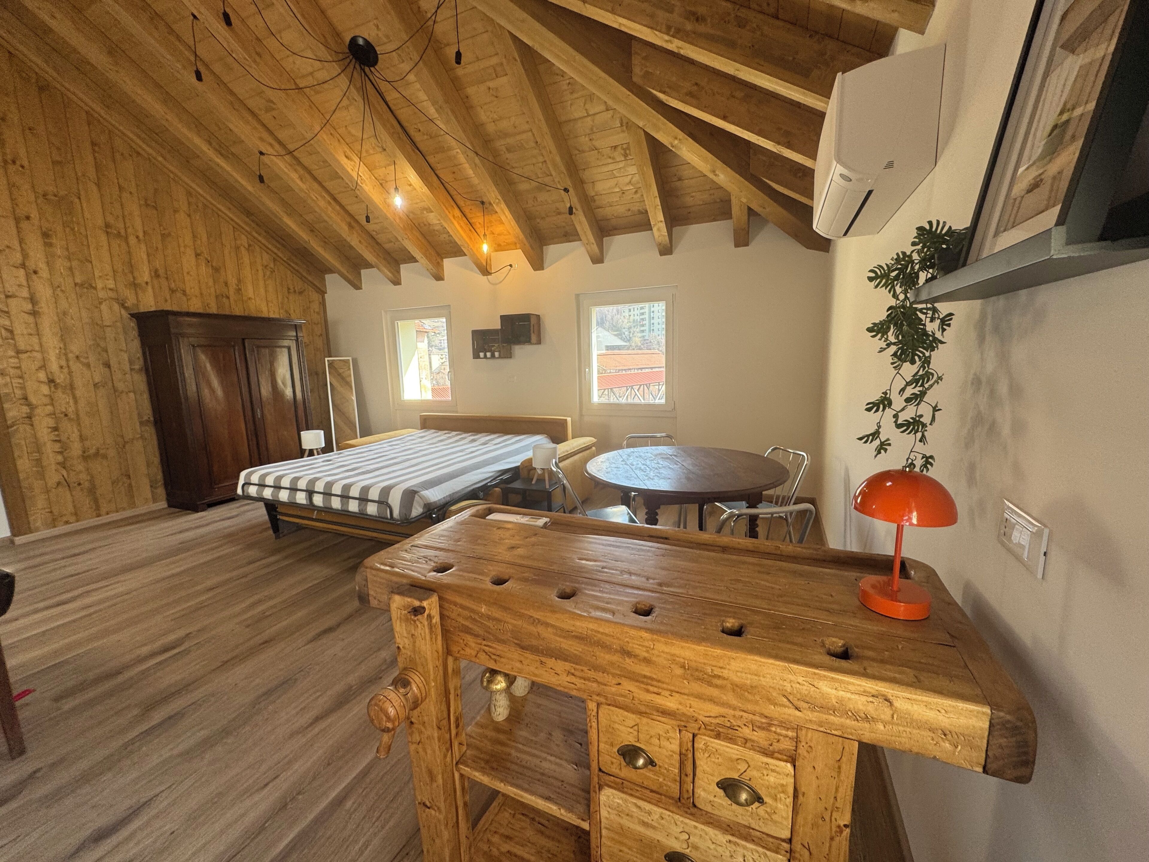 A room with a wooden ceiling, a bed, a table with chairs, a desk, and a window.