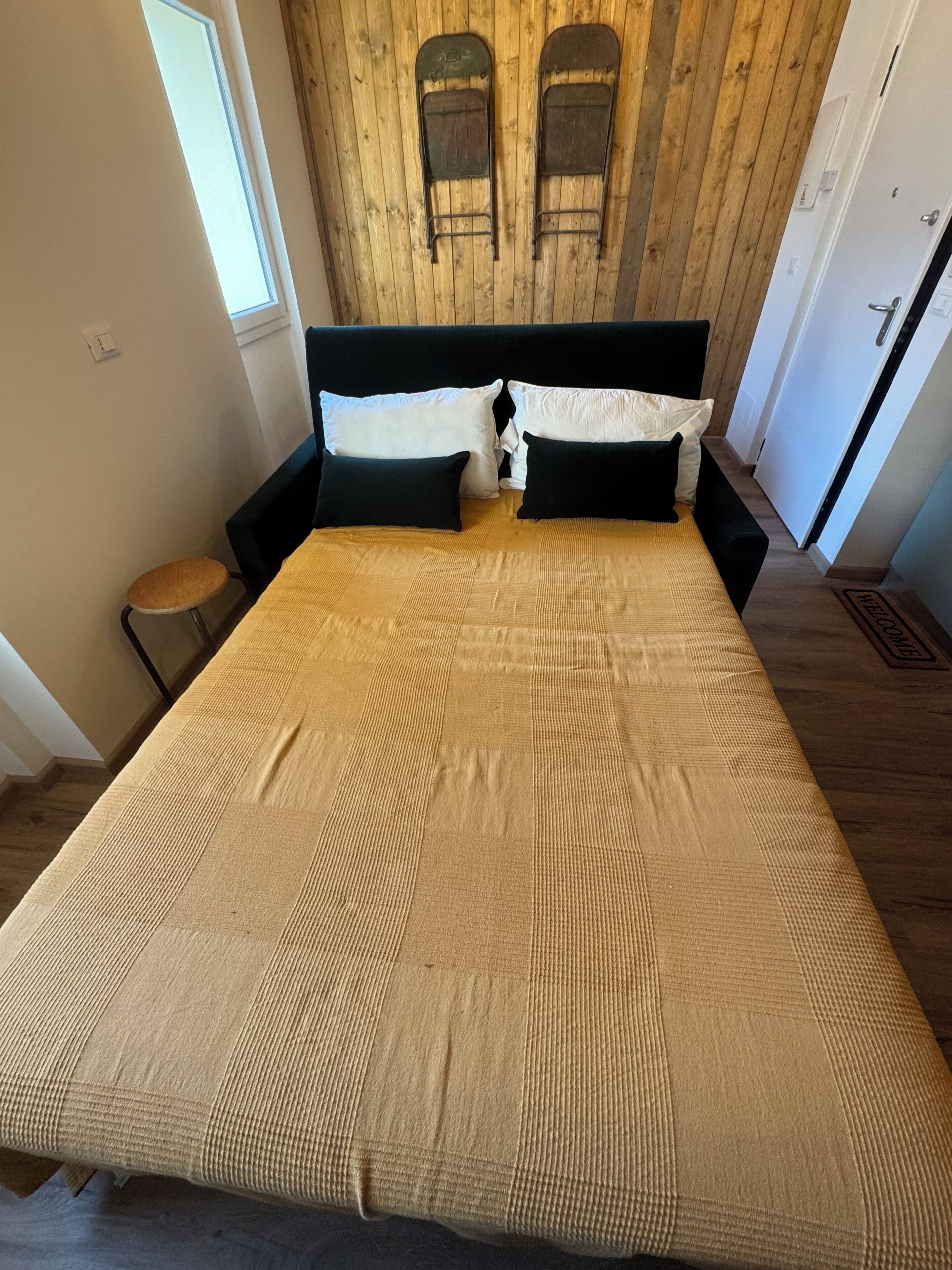 A bed with a beige bedspread, two black headboards, and a wooden wall with two small windows.