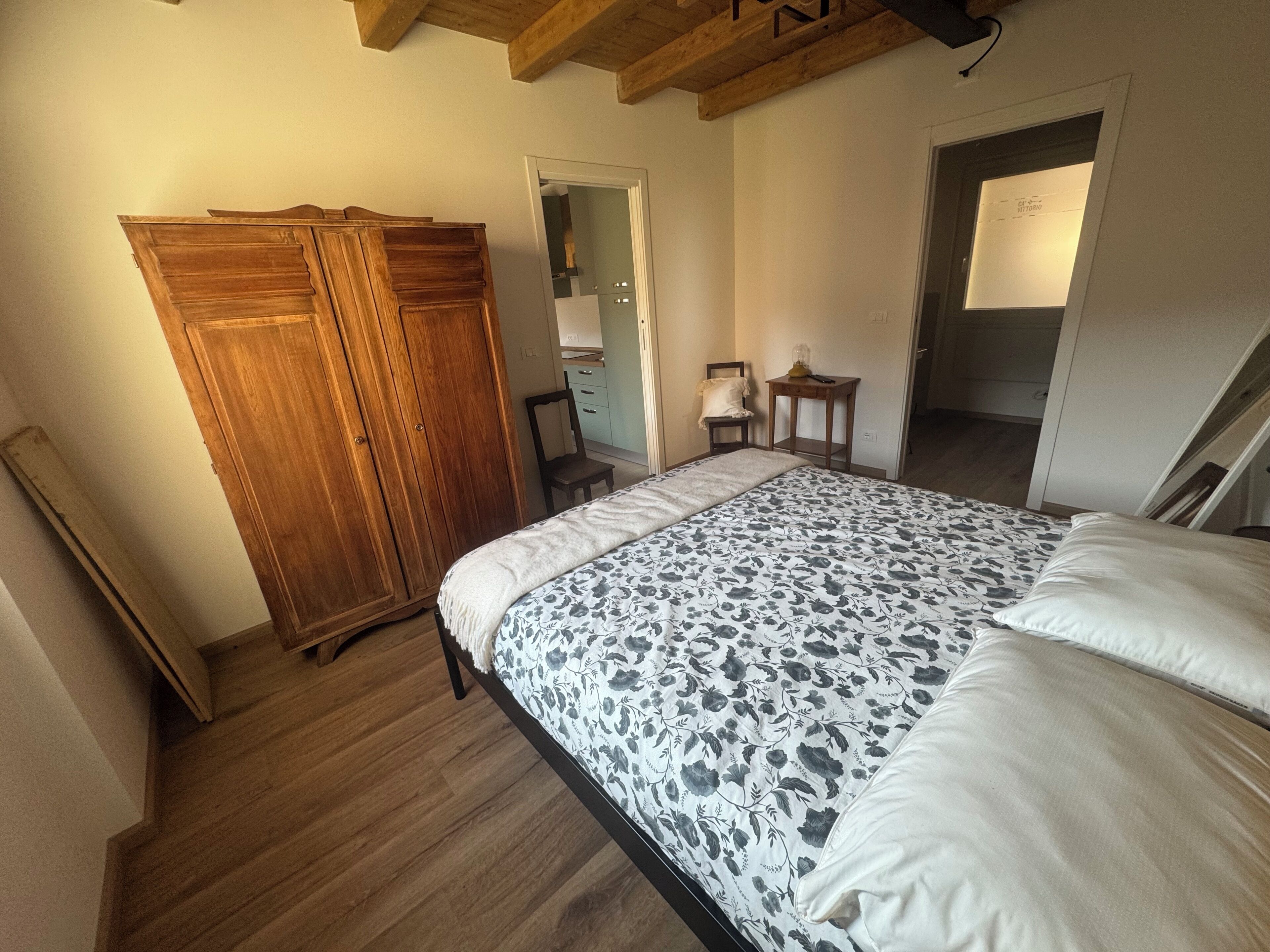 A bedroom with a wooden wardrobe, a bed with a floral bedspread, a small bedside table, and a chair.