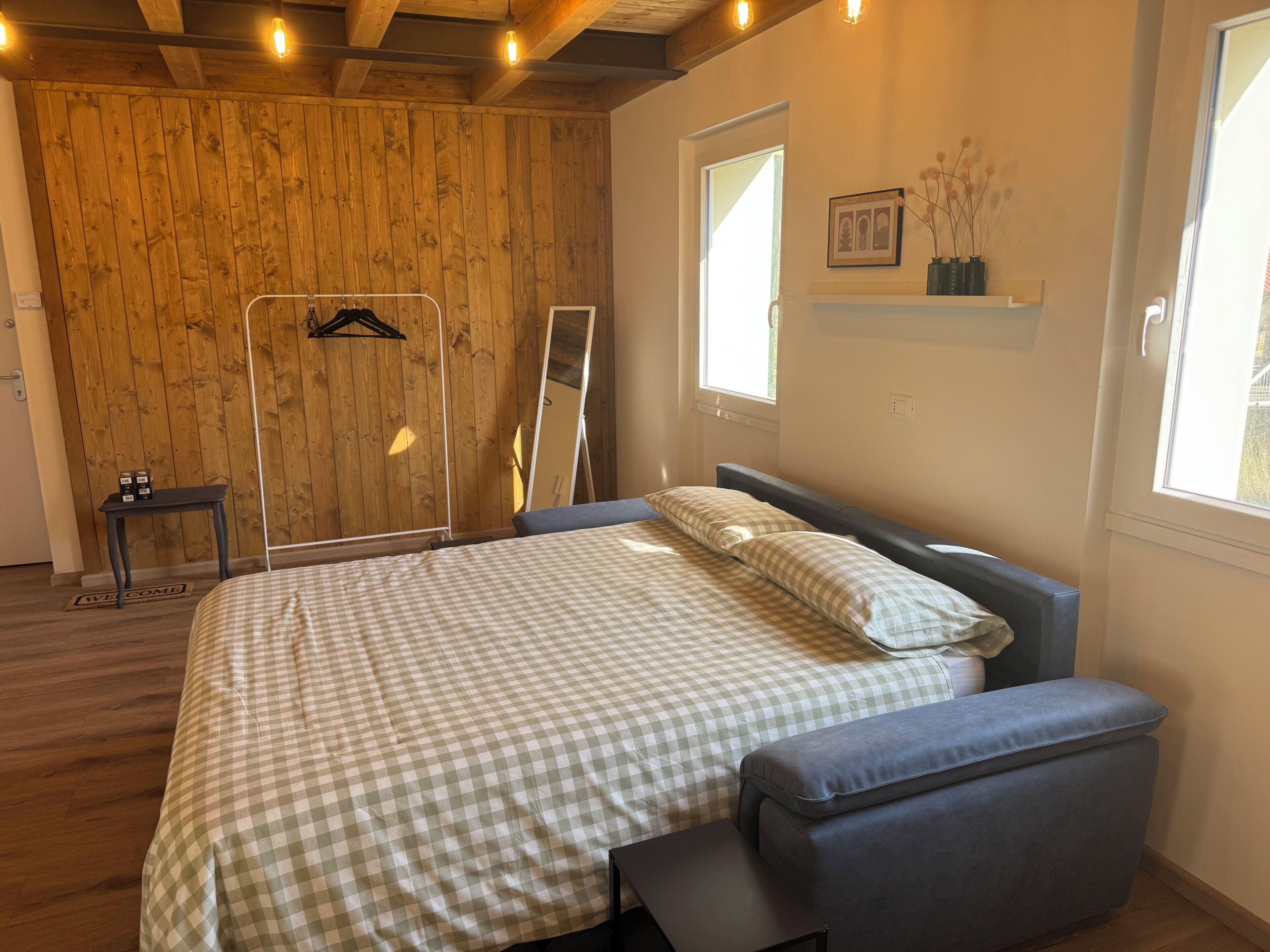 A bedroom with a checkered bedspread, a grey upholstered bench, a wooden wardrobe, and a window with a view.