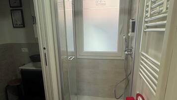 Une salle de douche fermée par des parois de verre, dotée d’une fenêtre dépolie, d’un seau rouge et d’un porte-serviettes.