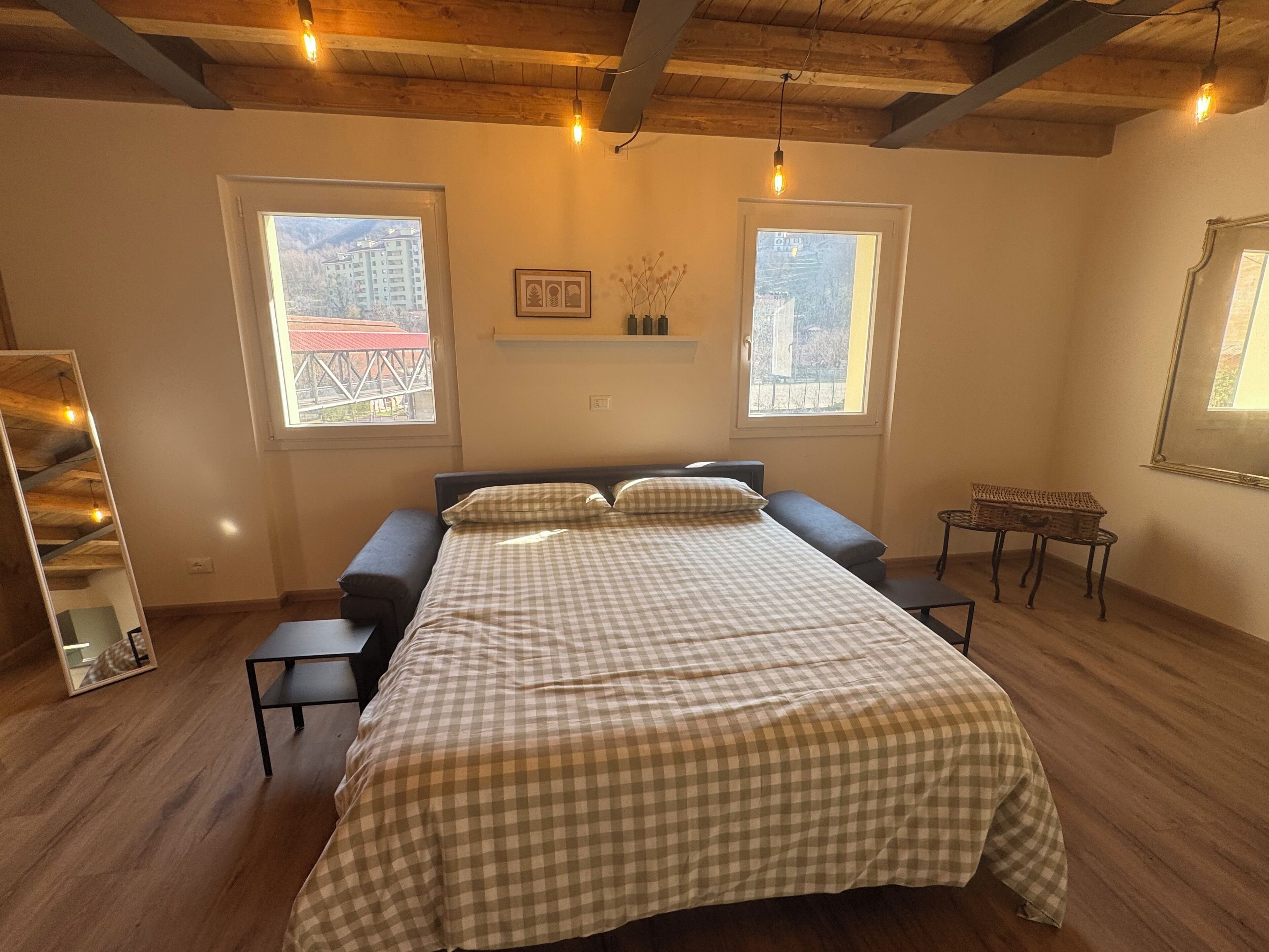 Un dormitorio con una cama, dos ventanas, techo de madera y una pequeña mesa con una cesta.