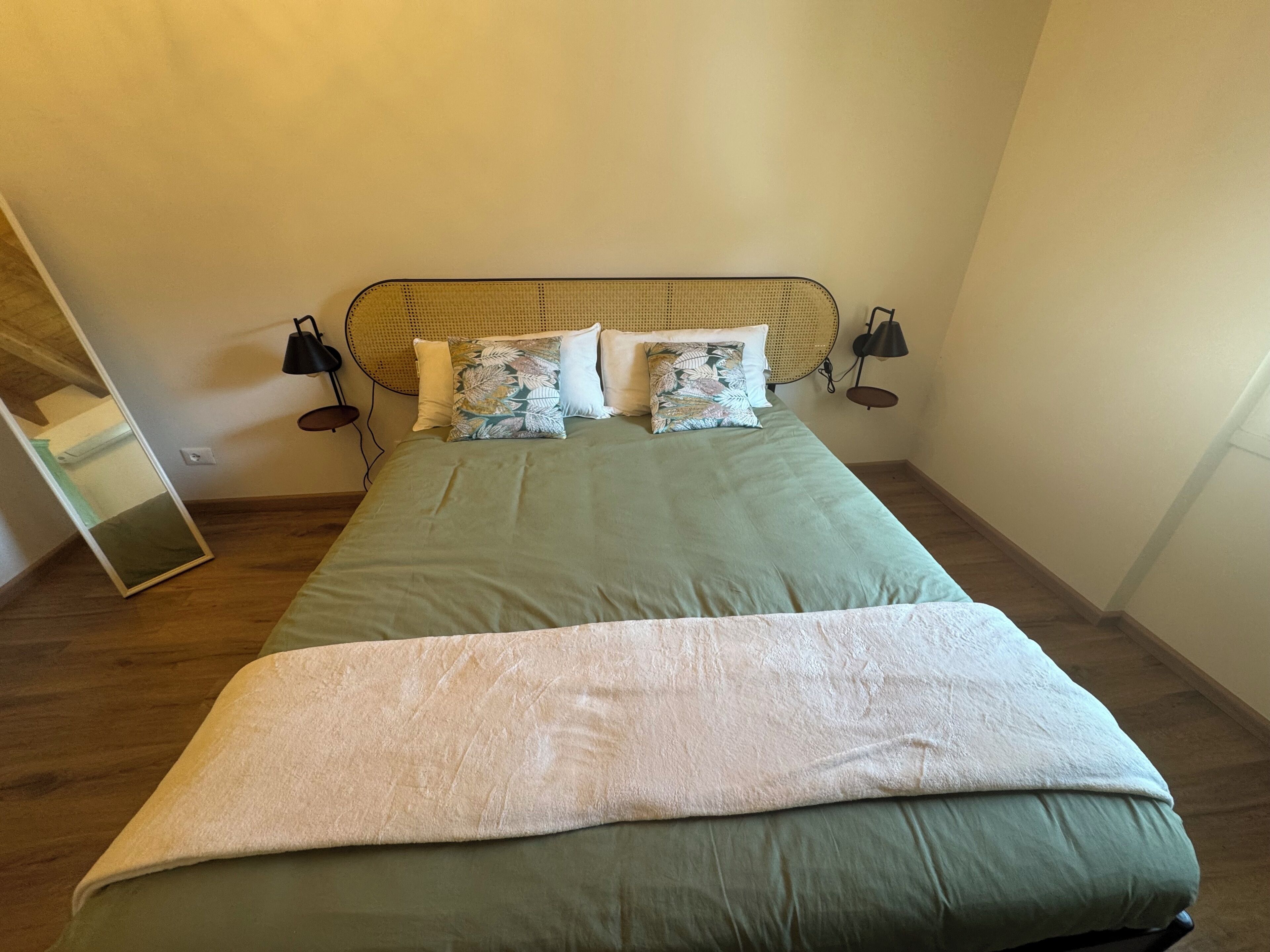 A bed with a green bedspread, a white throw blanket, and multiple pillows, including a floral one. There are two bedside lamps and a mirror on the wall.