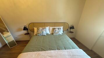A bed with a green bedspread, a white throw blanket, and multiple pillows, including a floral one. There are two bedside lamps and a mirror on the wall.
