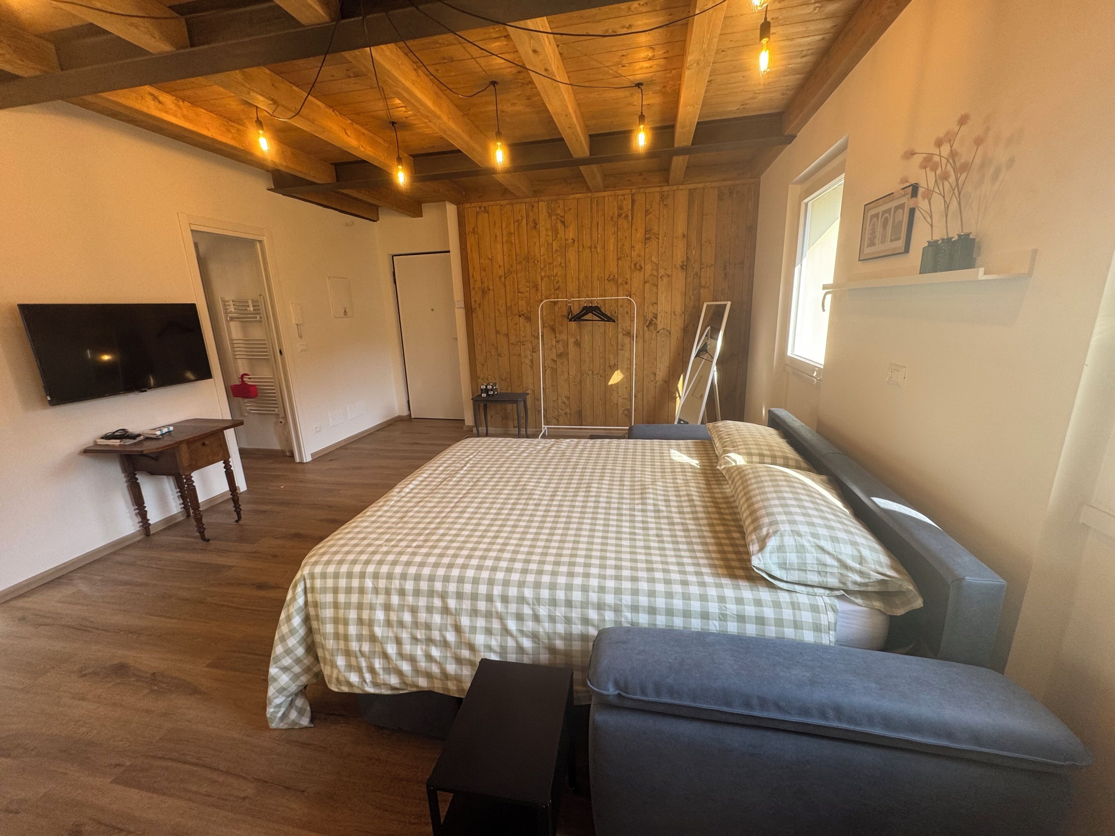 A bedroom with a wooden ceiling, a bed with checkered bedding, a bedside table, a bench, a TV, and a window with a picture frame.