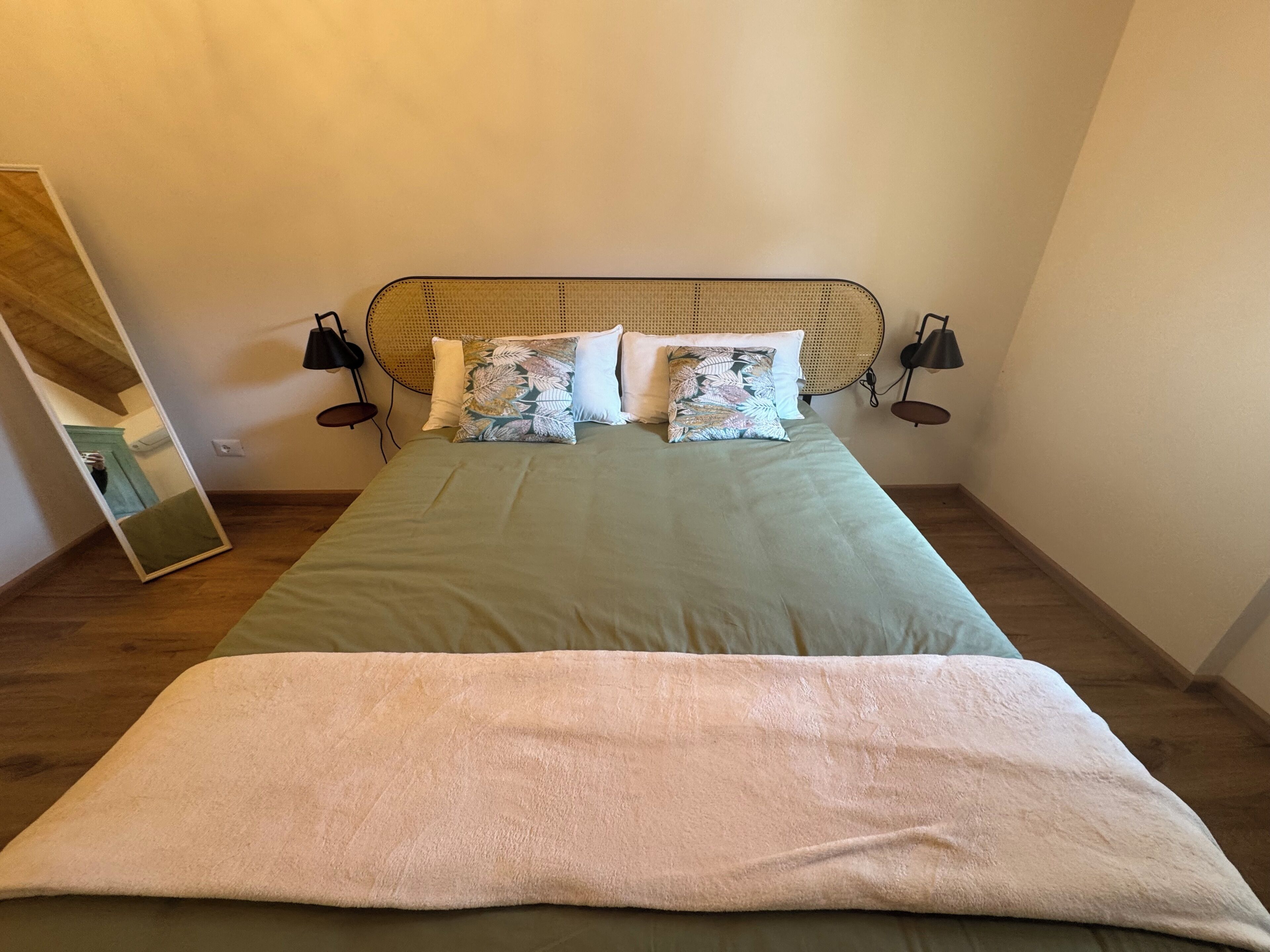 A bed with a green bedspread, two patterned pillows, a white throw blanket, and a rattan headboard.