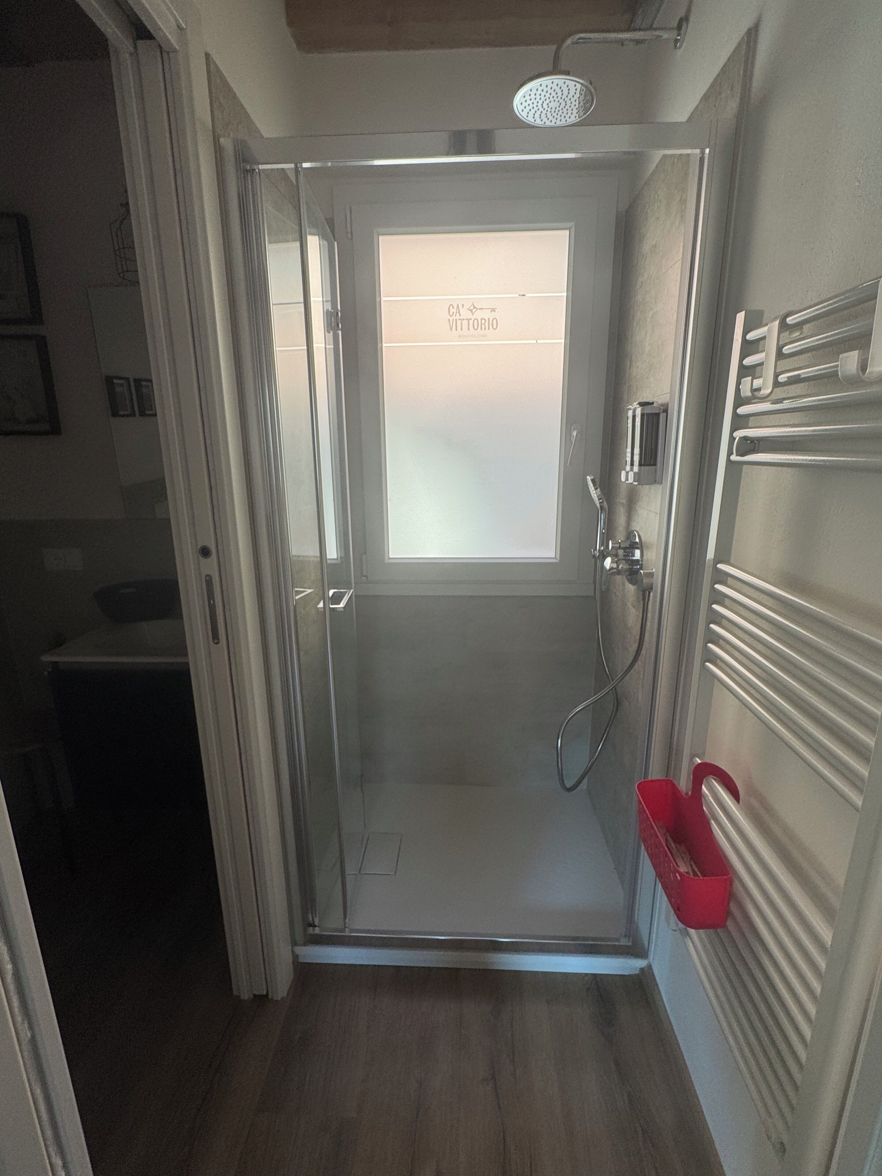 Ducha con cerramiento de vidrio y puerta esmerilada, un cubo rojo y un perchero en la pared.