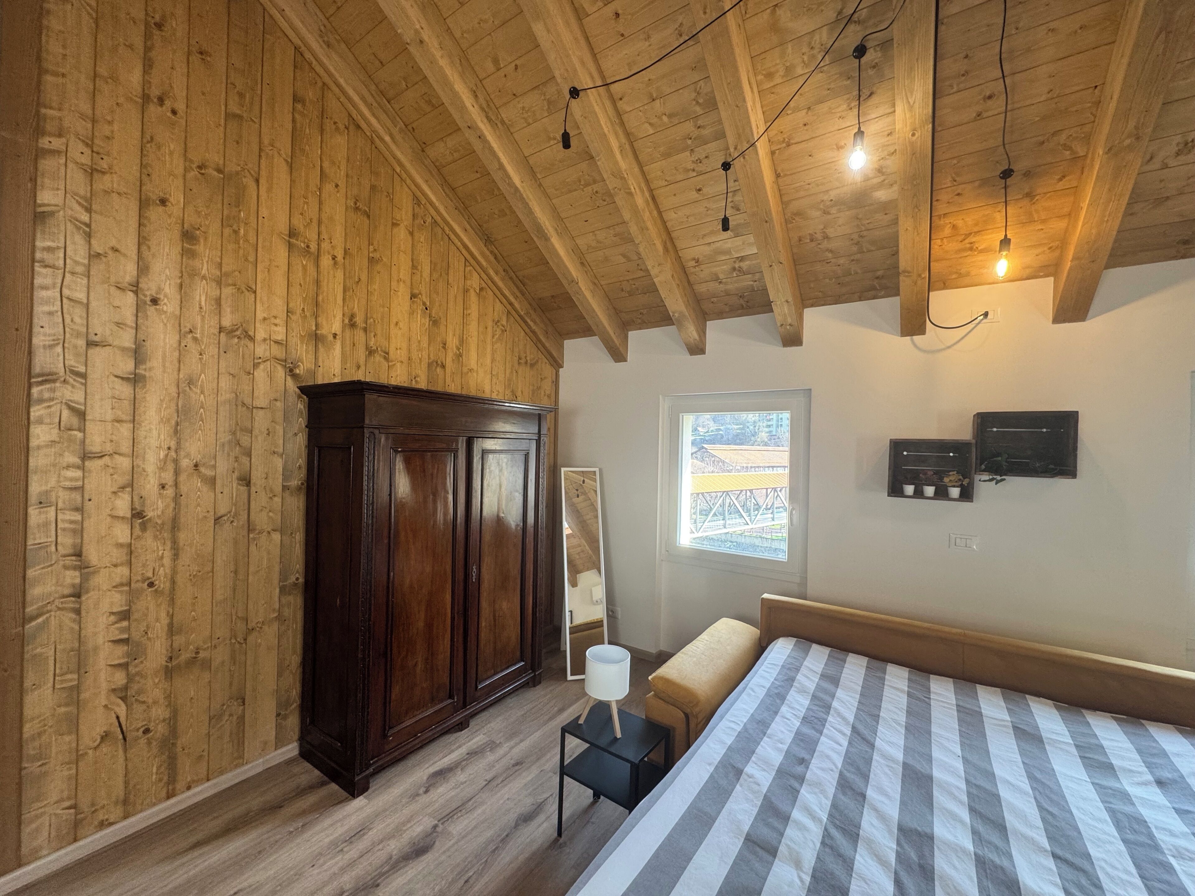 Une chambre avec des murs en bois, un grand lit, une armoire, une petite table avec une lampe et une fenêtre donnant sur un beau paysage.