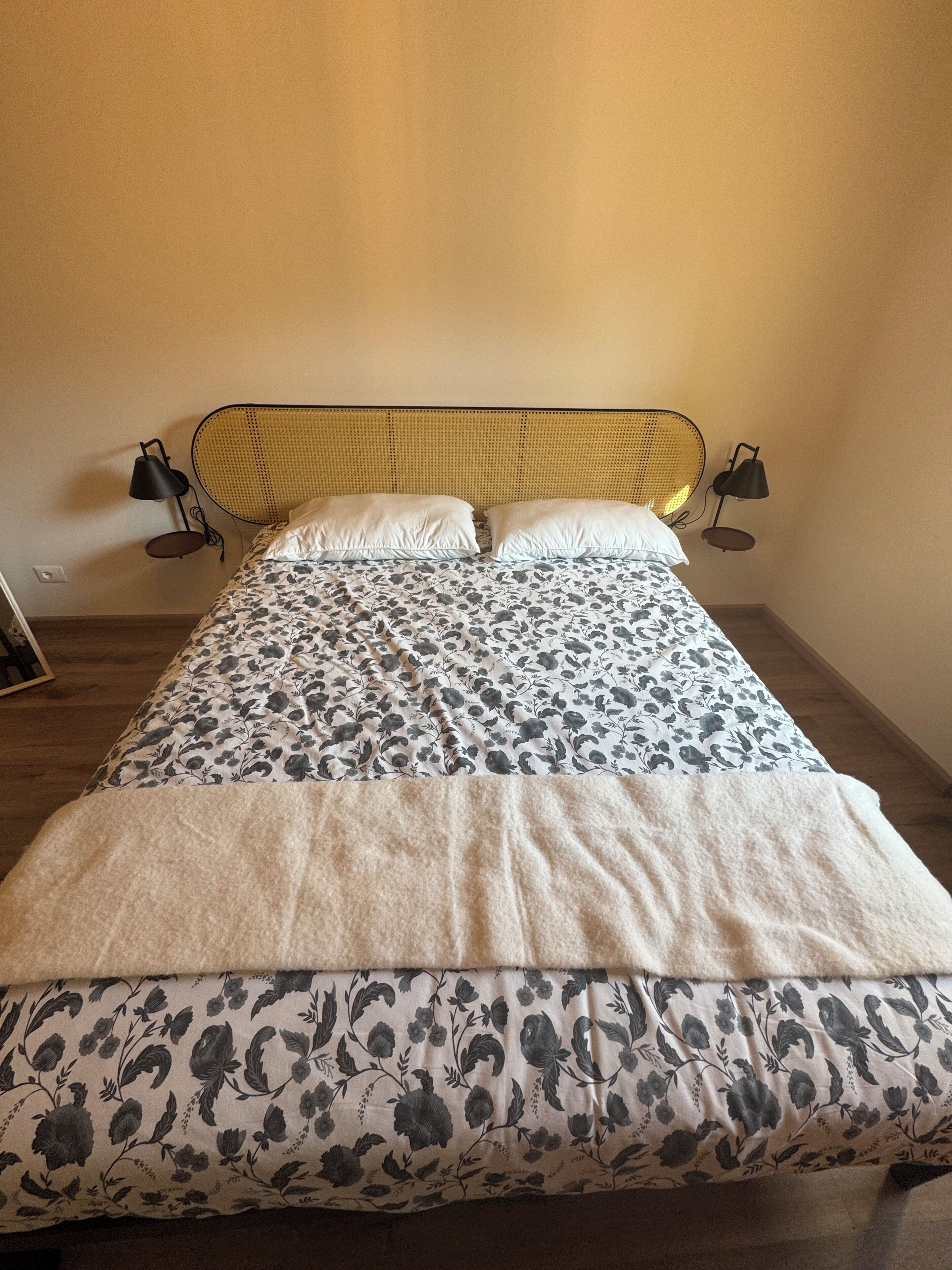 Cama com cabeceira trançada, duas luminárias de cabeceira e edredom estampado com motivos florais.