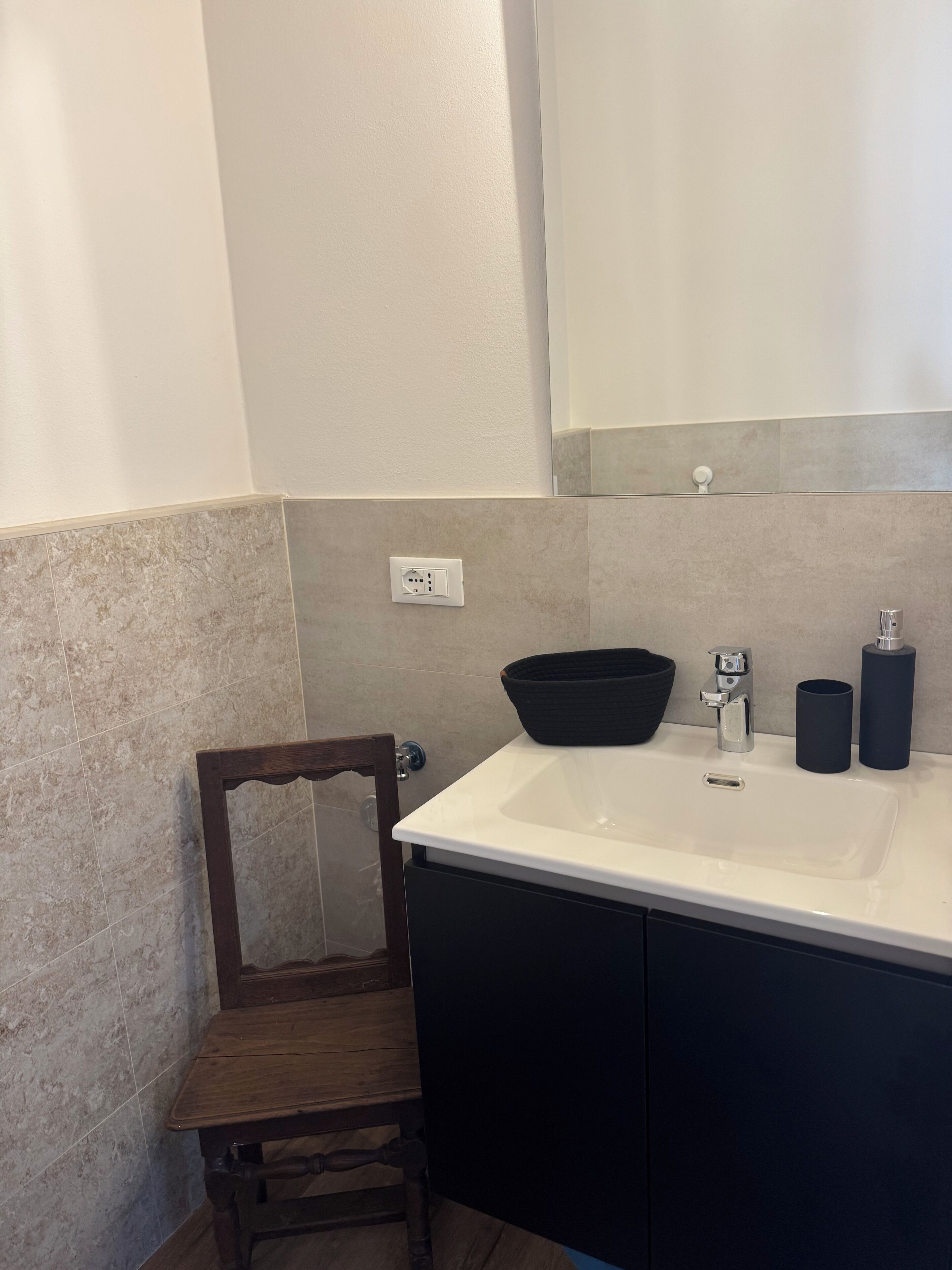 A bathroom with a white sink, dark cabinets, a wooden chair, and a mirror above the sink.