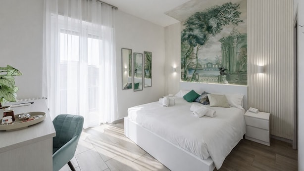 Ett modernt hotellrum med en stor säng, ett skrivbord med en stol, ett stort fönster med tunna gardiner och en väggmålning på väggen.